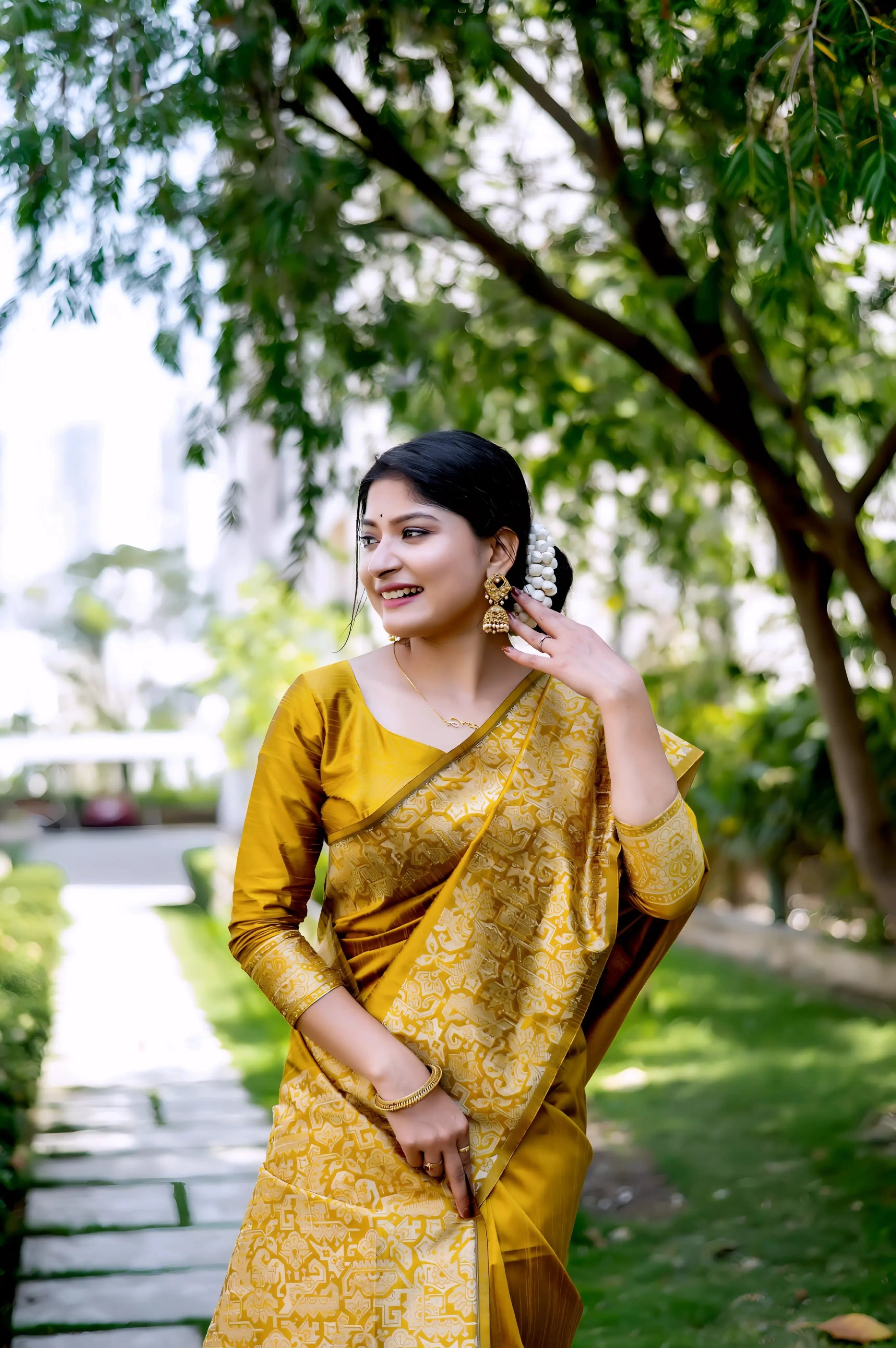 A woman models a luxurious Golden Yellow Raw Silk Saree. The saree features a wide border and pallu with dense, tone-on-tone gold Zari weaving in a Kalamkari-style floral and paisley pattern. She wears a matching long-sleeve yellow blouse and traditional gold jewelry, including a maang tikka, posing outdoors on a sunlit garden path.